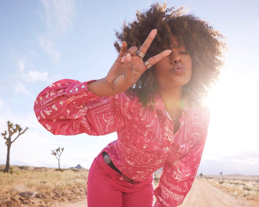 Woman in pink western shirt and pants making a peace sign in desert landscape under blue sky with Joshua trees in the background.