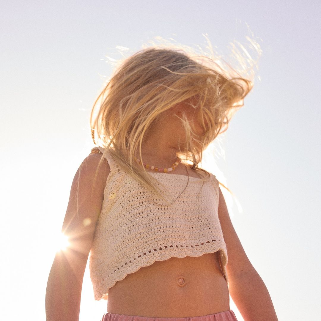 Young child stands in bright sunlight wearing a white crocheted crop top and pink shorts, with wind blowing through blonde hair.