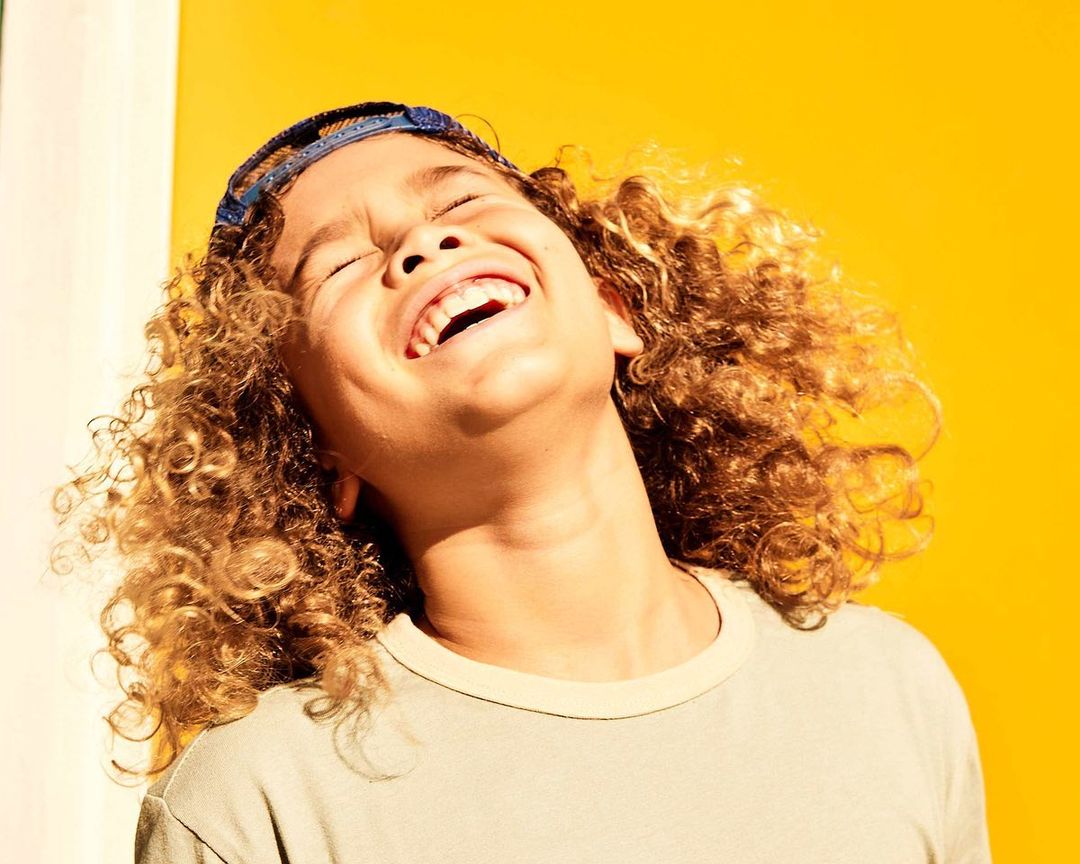 A child with curly hair, wearing a headband and grey shirt, laughs with their head tilted back against a yellow and white background.