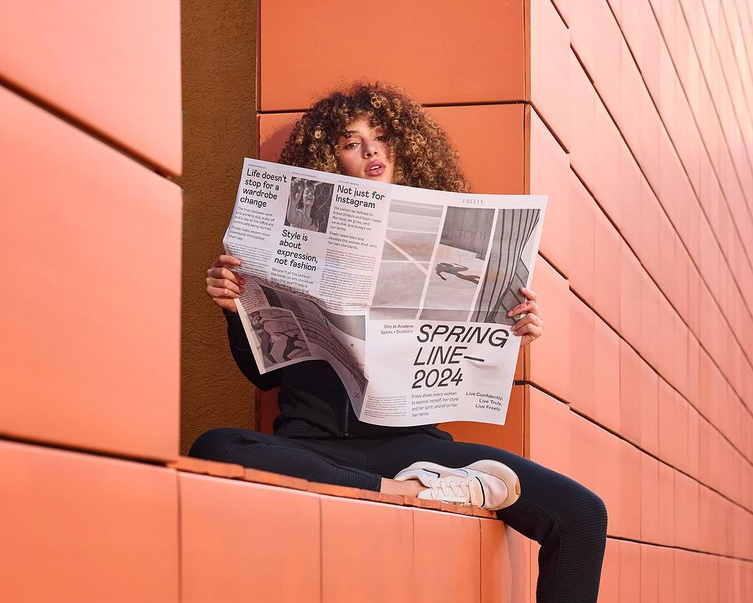 A young adult female model with curly hair sits outdoors in stylish athletic apparel, reading a newspaper in front of an orange wall.