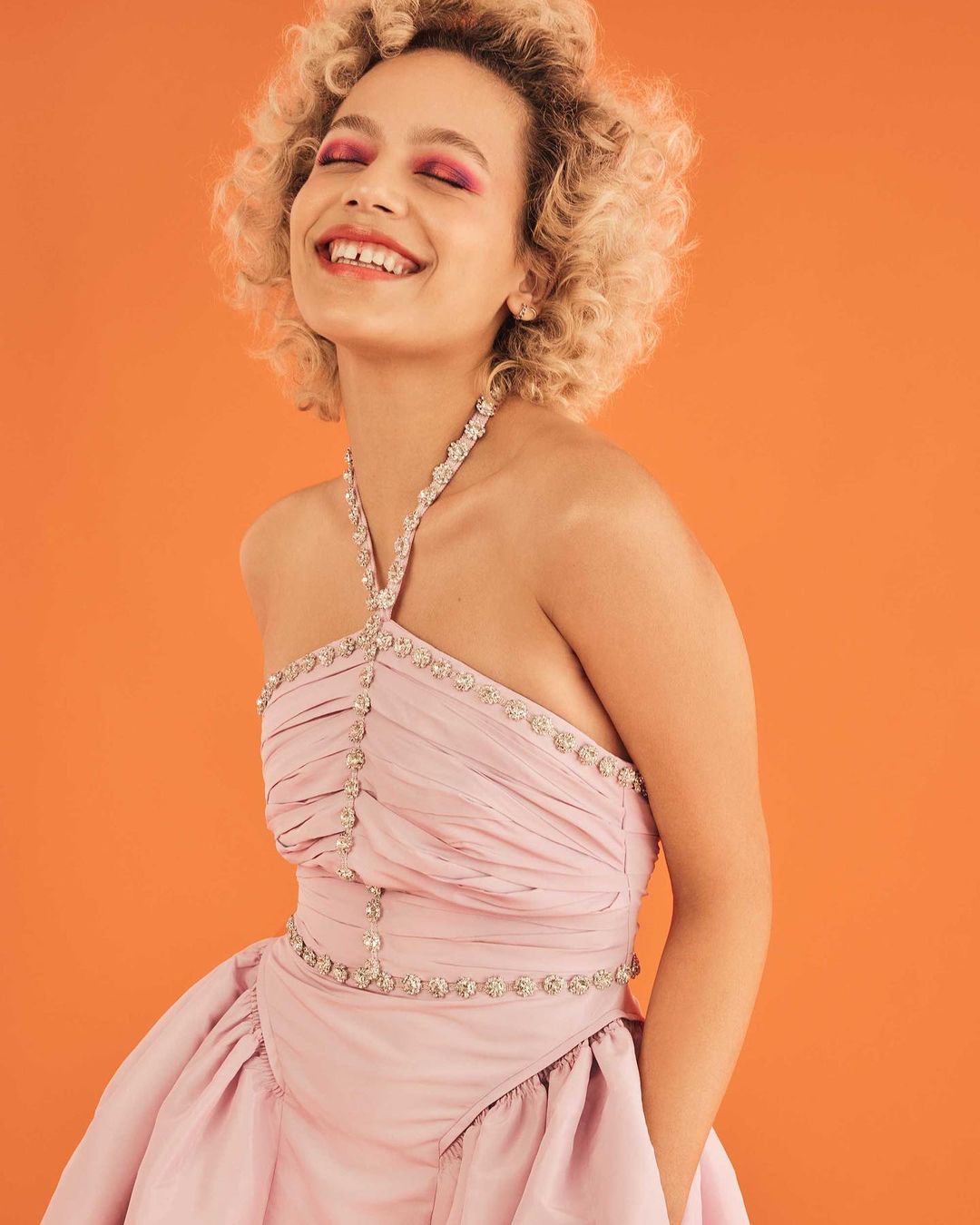 Smiling woman with curly hair and bright pink makeup poses in a pink gown against a vibrant orange backdrop in a studio setting.