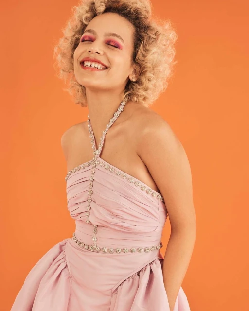 Smiling woman with curly hair and bright pink makeup poses in a pink gown against a vibrant orange backdrop in a studio setting.