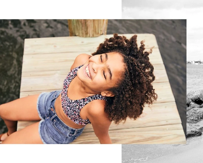 Young girl smiles with eyes closed, laying on a wooden dock wearing a floral bathing suit top and denim shorts near the ocean.