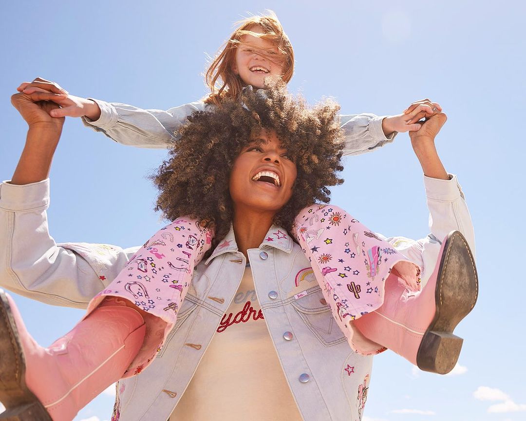 Mother and daughter share a happy moment outdoors with a blue sky backdrop. The daughter rides on her mother's shoulders, both smiling.