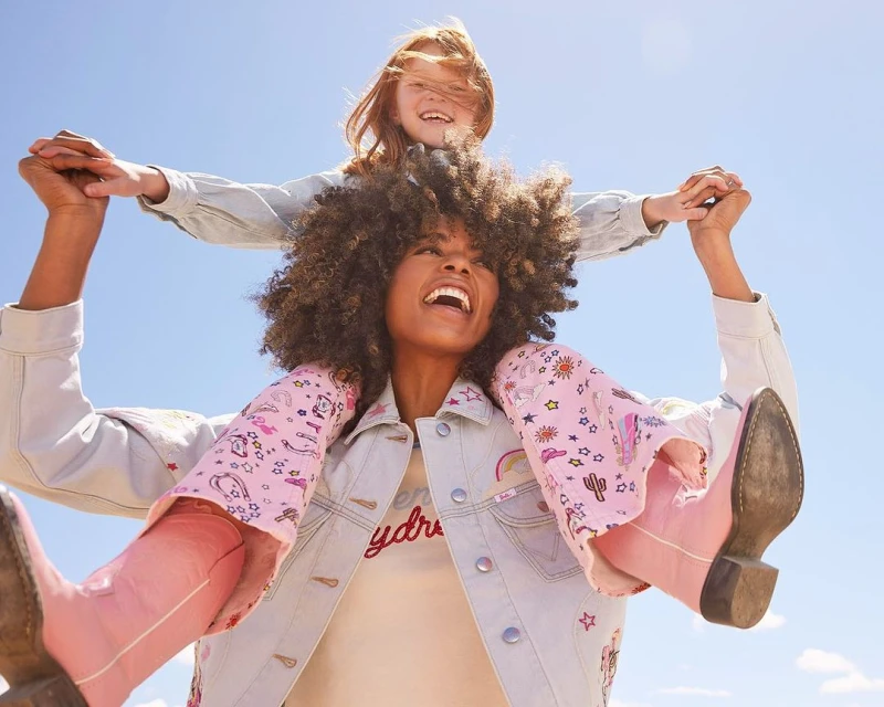 Mother and daughter share a happy moment outdoors with a blue sky backdrop. The daughter rides on her mother's shoulders, both smiling.