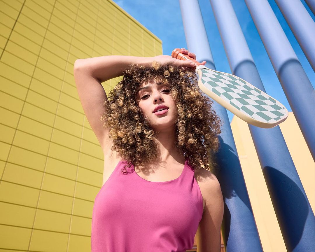 Woman posing with paddle ball racket in front of colourful striped building, bright sunny day, close-up shot, pink athletic top.