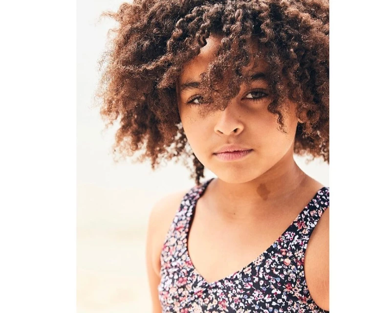 Close-up portrait of a beautiful child with curly brown hair, wearing a floral-patterned top against a blurred light background outdoors.