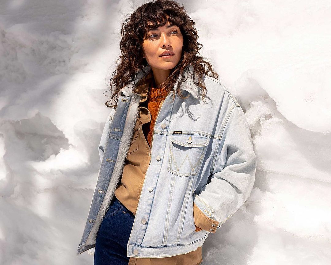 Brunette woman wearing a denim sherpa jacket and tan shirt poses against a cloud-like background in a medium studio shot.