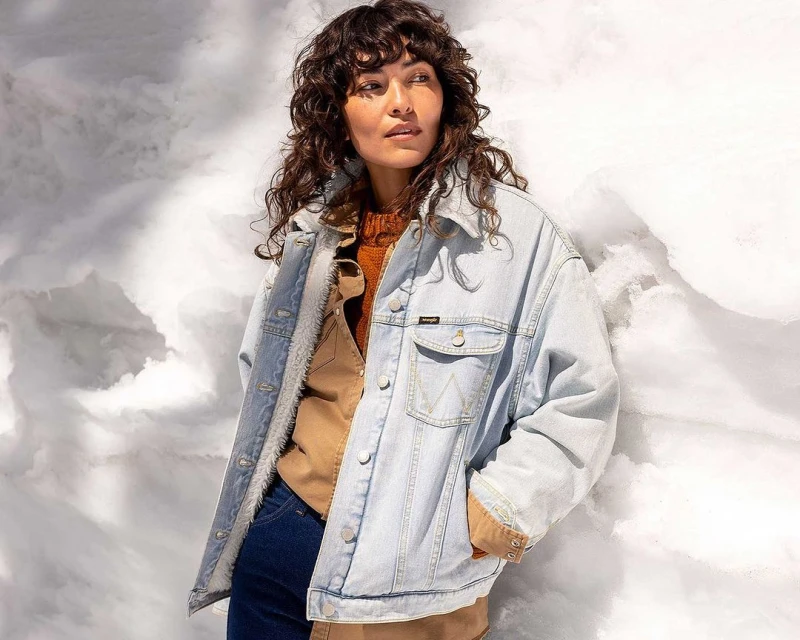 Brunette woman wearing a denim sherpa jacket and tan shirt poses against a cloud-like background in a medium studio shot.