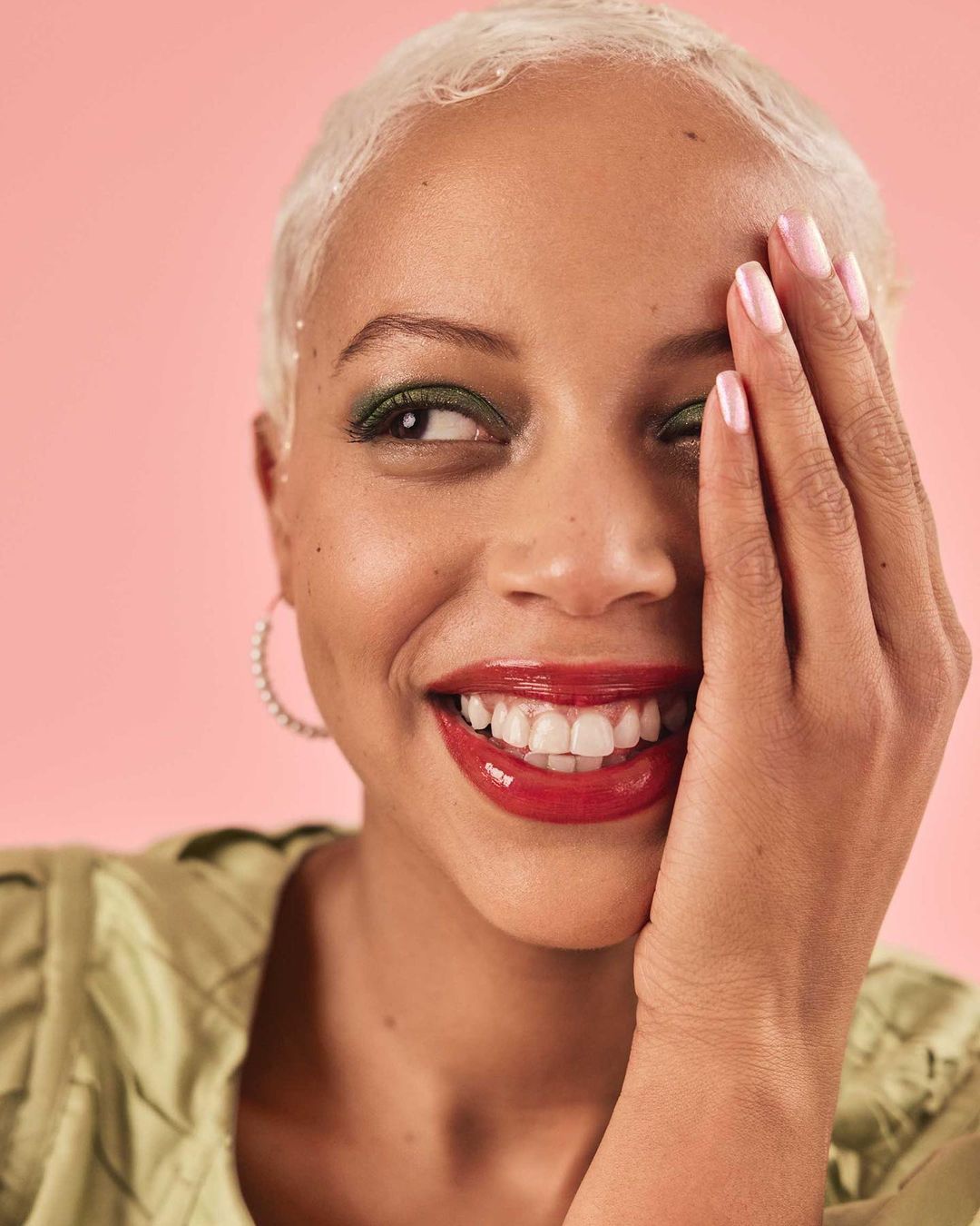Close-up studio shot of a smiling young adult woman with short white hair and green eyeshadow, wearing a light green top against pink.