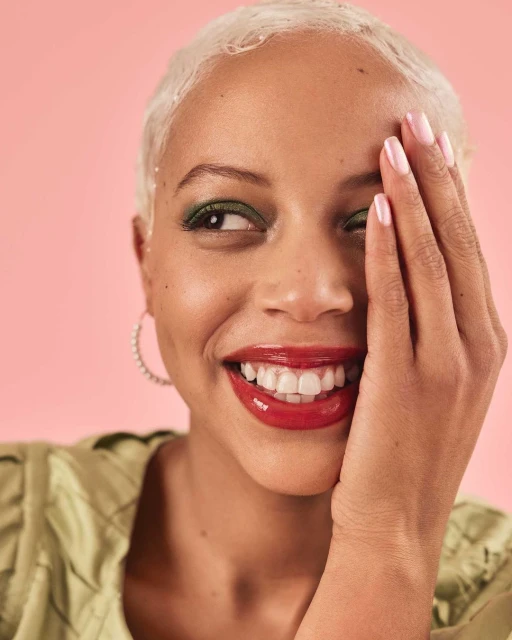 Close-up studio shot of a smiling young adult woman with short white hair and green eyeshadow, wearing a light green top against pink.