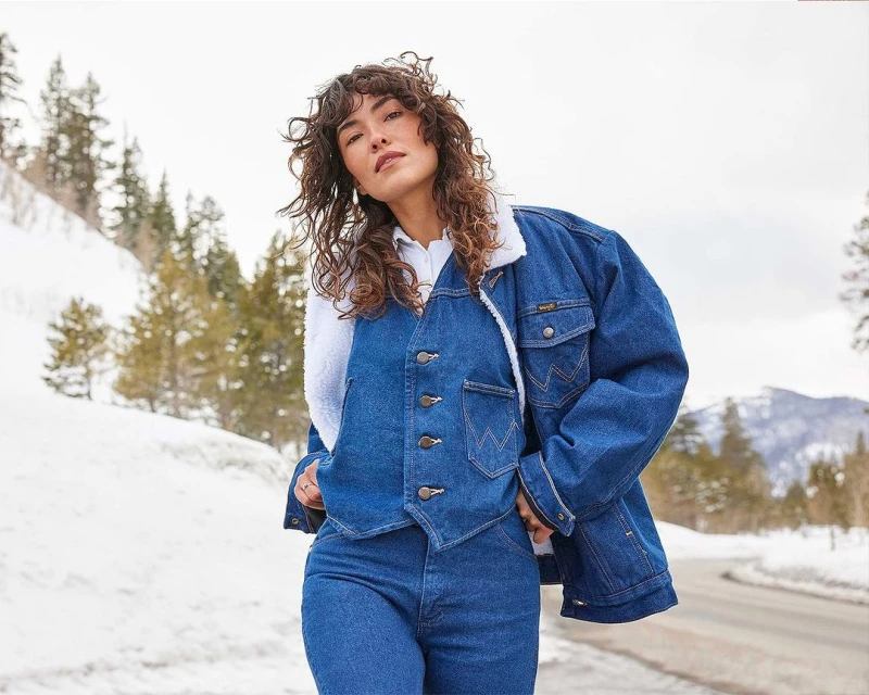 Woman in denim jacket and jeans poses on snowy road near snow-covered trees and mountains against cloudy sky, outdoor fashion.