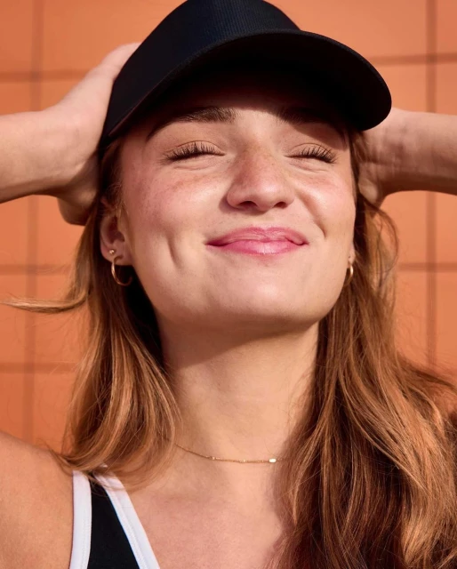 Portrait of a young woman with freckles wearing a black visor and gold jewelry smiling in front of a textured orange wall.