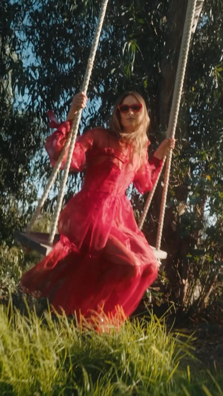 A woman in a red dress swings under a tree. The image is shot at ground level with shallow focus on the woman and swing.