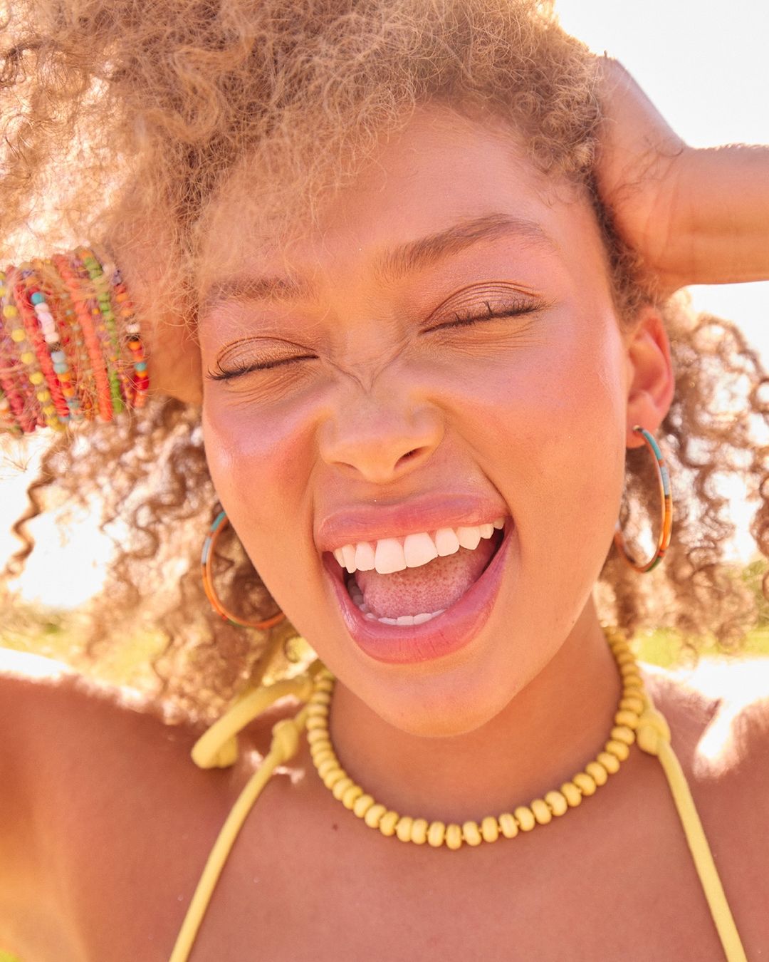 Close-up of a smiling young woman with curly blonde hair wearing a yellow beaded necklace and hoop earrings, with green nature background.