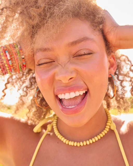Close-up of a smiling young woman with curly blonde hair wearing a yellow beaded necklace and hoop earrings, with green nature background.