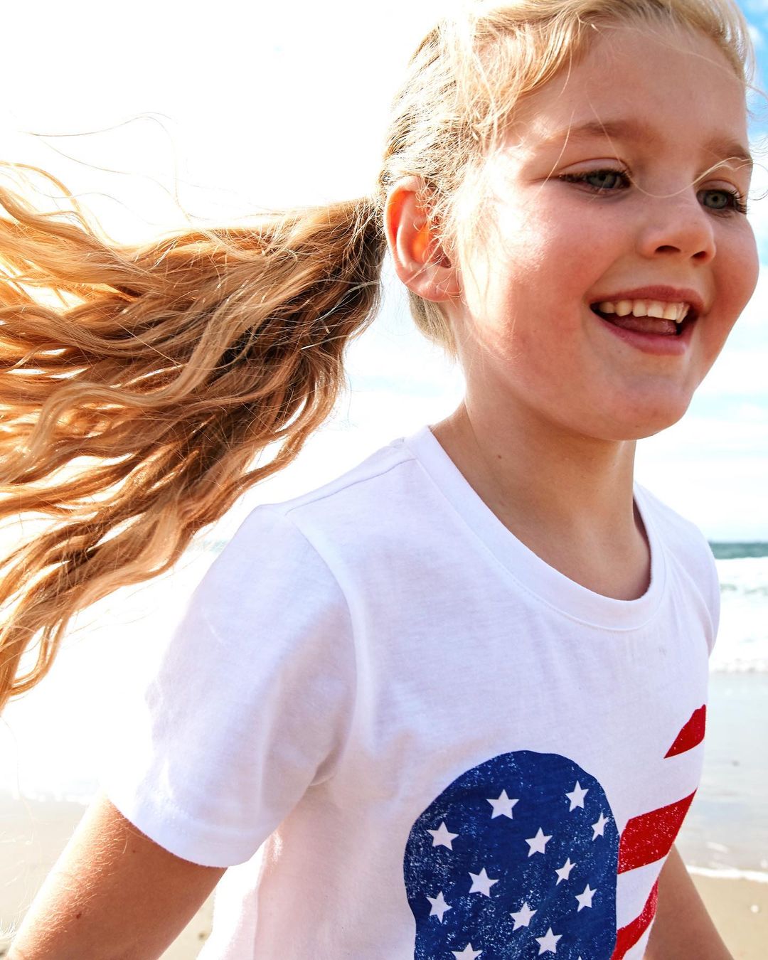 A blonde child with hair in a ponytail smiles broadly at a beach while wearing a t-shirt with a patriotic American flag design.