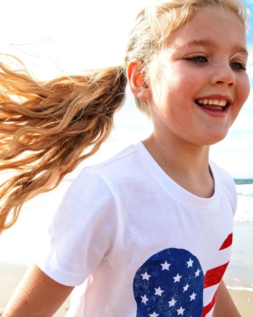 A blonde child with hair in a ponytail smiles broadly at a beach while wearing a t-shirt with a patriotic American flag design.