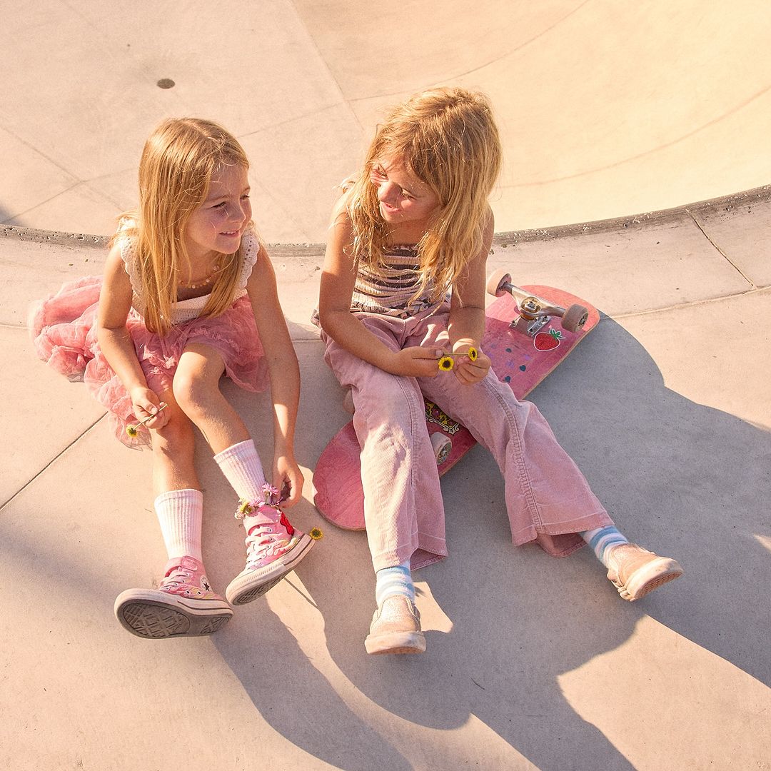 Two young girls sit on a pink skateboard at a skatepark, smiling at each other, wearing pink outfits and high-top sneakers in soft, natural light.