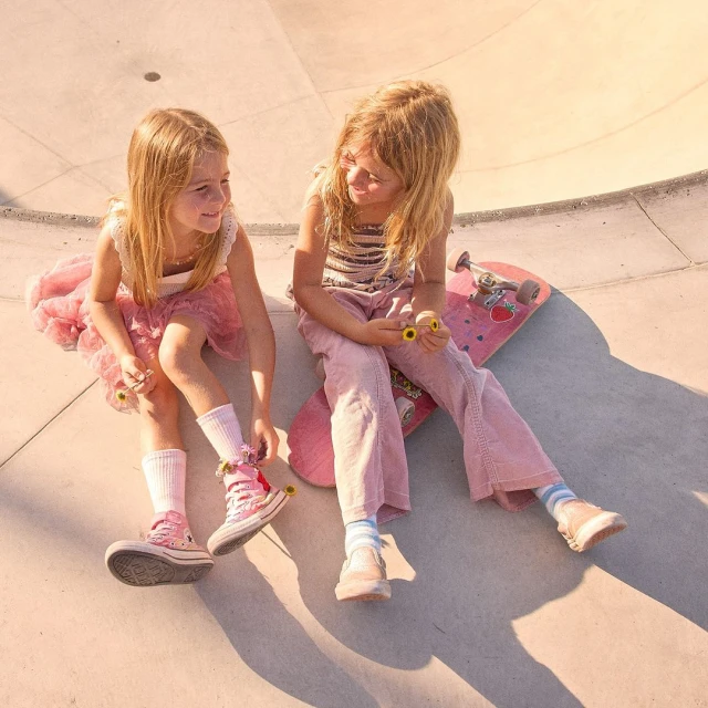 Two young girls sit on a pink skateboard at a skatepark, smiling at each other, wearing pink outfits and high-top sneakers in soft, natural light.