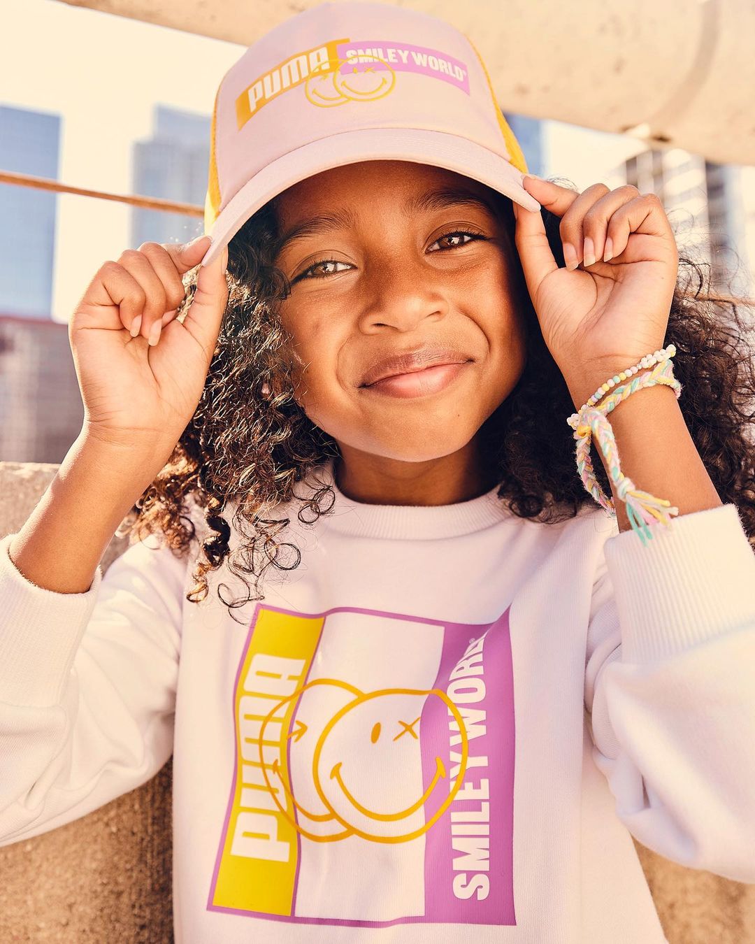 Portrait of a young girl wearing a Puma-branded hat and sweatshirt with a Smiley World graphic, smiling outdoors in daylight.