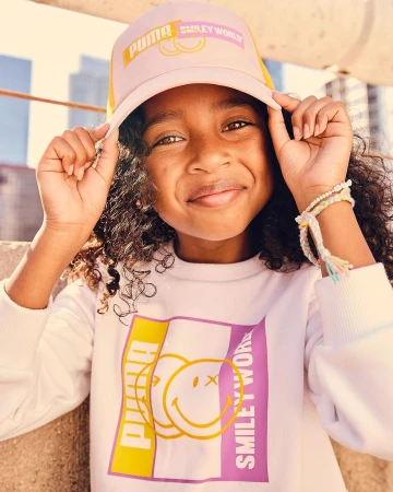 Portrait of a young girl wearing a Puma-branded hat and sweatshirt with a Smiley World graphic, smiling outdoors in daylight.