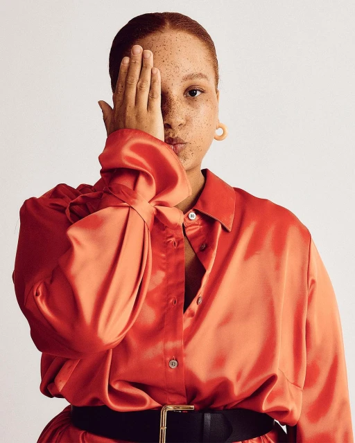 Red-haired woman in an orange satin shirt and black belt covers one eye with her hand in a studio portrait against a white background.