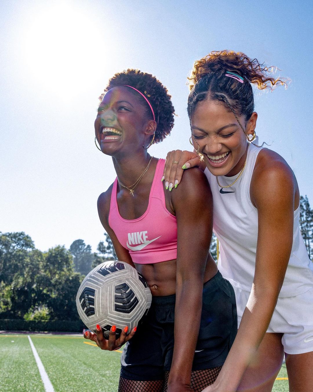Two smiling women, one holding a soccer ball, stand together on a green soccer field under a clear blue sky in an eye-level shot.
