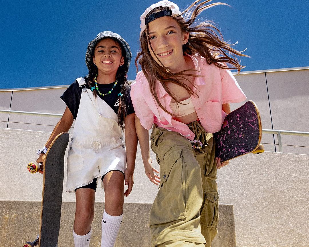Two smiling girls, one with a skateboard, are outdoors on a sunny day, wearing fashionable streetwear clothing. They are looking directly at the camera.
