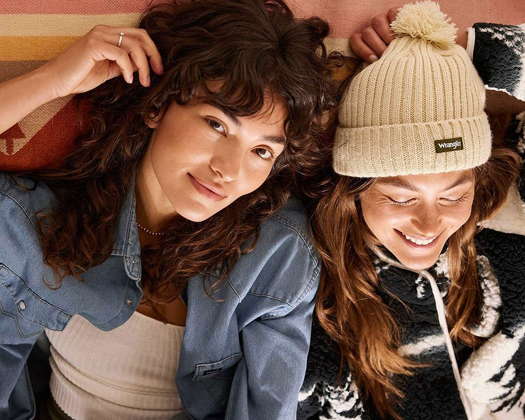 Two women wearing hats and denim jacket smiling, laying on a blanket with geometric design, eye-level angle, indoor setting.