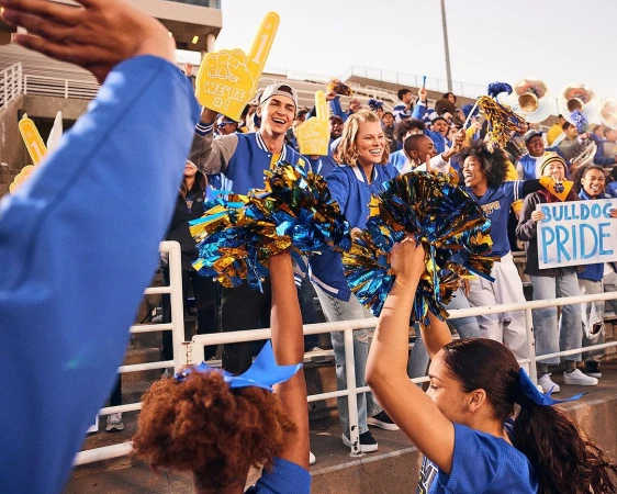 Enthusiastic crowd of football fans and cheerleaders in blue and gold team colors at an outdoor stadium on a sunny day.