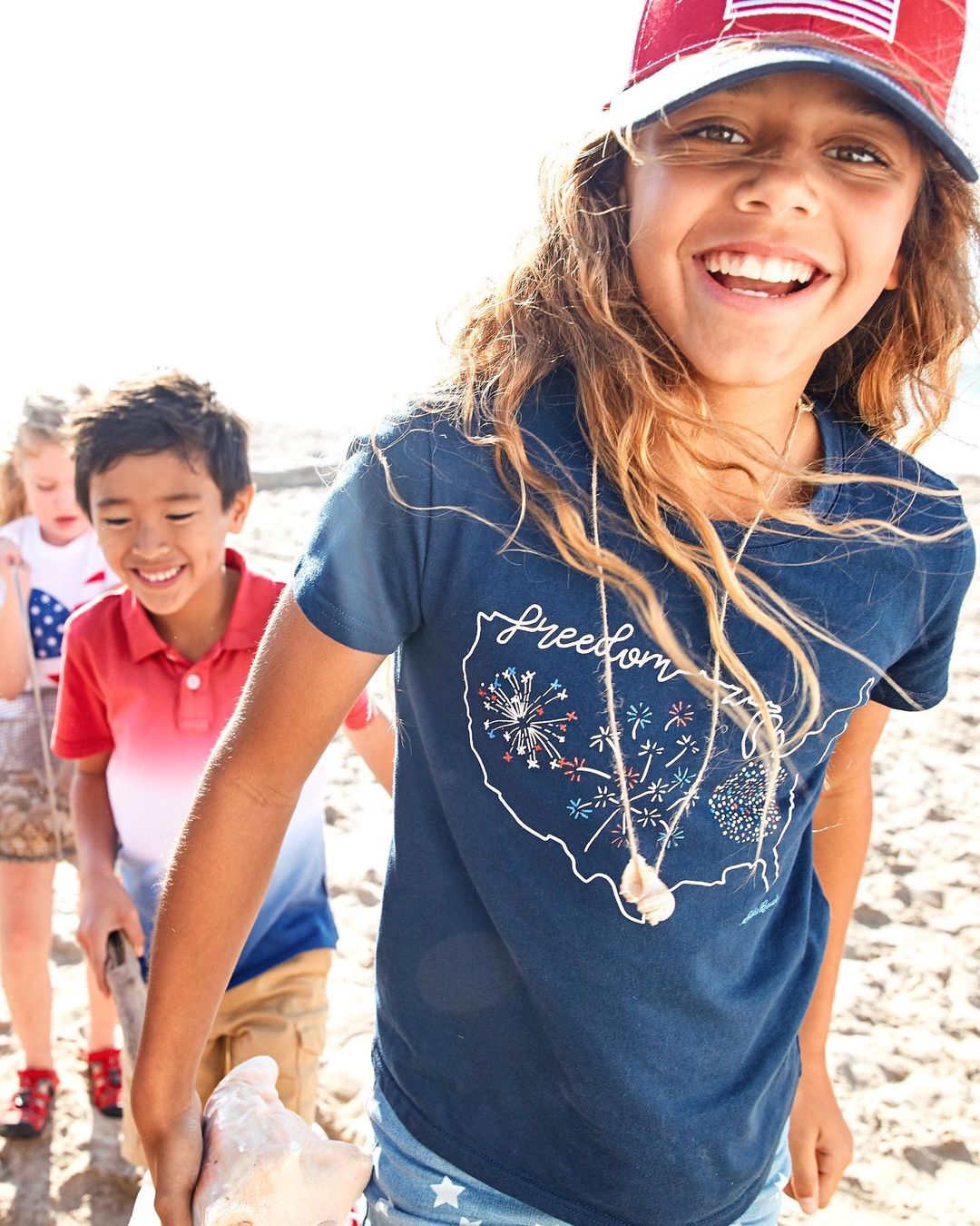 A group of children, including a girl wearing a patriotic t-shirt and cap, are seen at the beach, smiling and enjoying a sunny day.