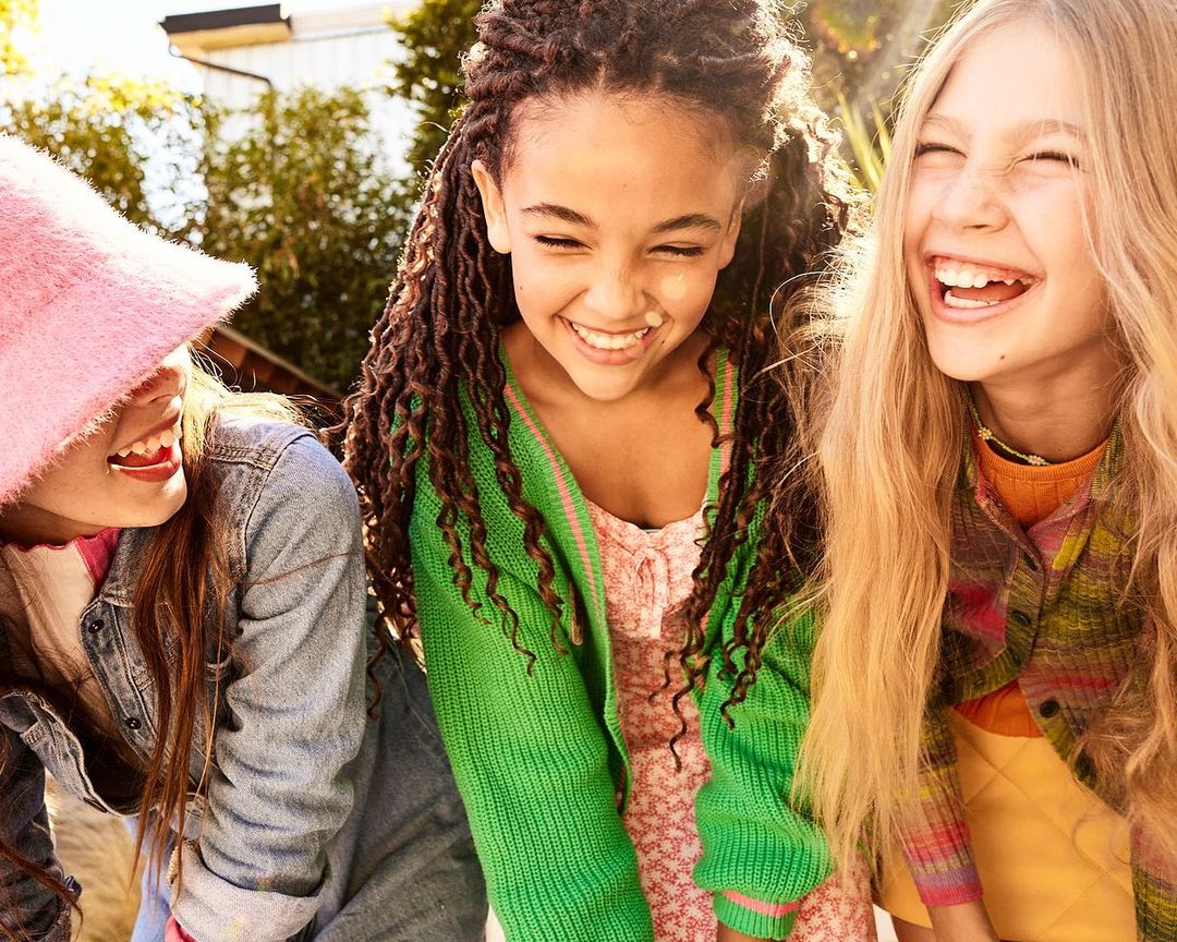 Three young girls outdoors, laughing together in bright sunshine. One wears a pink hat, another a green sweater, all in casual clothing.