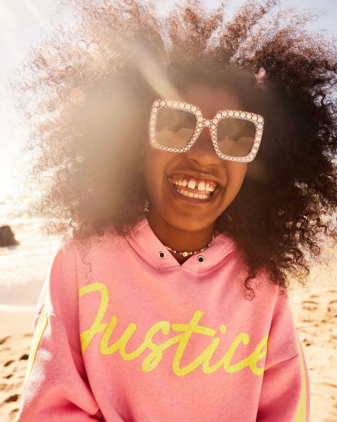 Smiling young Black girl with large afro hair, stylish sunglasses, and pink hoodie with a yellow logo, posing outdoors at the beach.