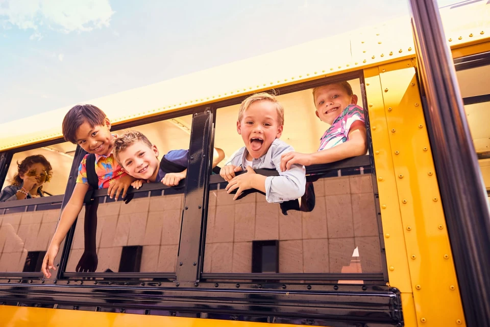 Group of smiling children looking out the windows of a yellow school bus. The windows are open as they look out.