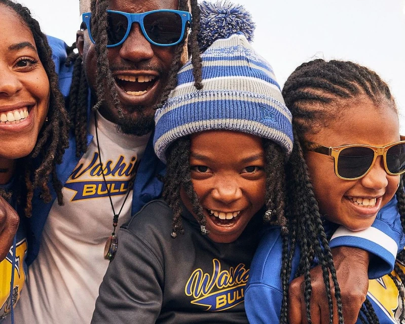 Four smiling people, two adults and two children, wearing blue and gold apparel. They all have dark hair and are smiling at the camera.