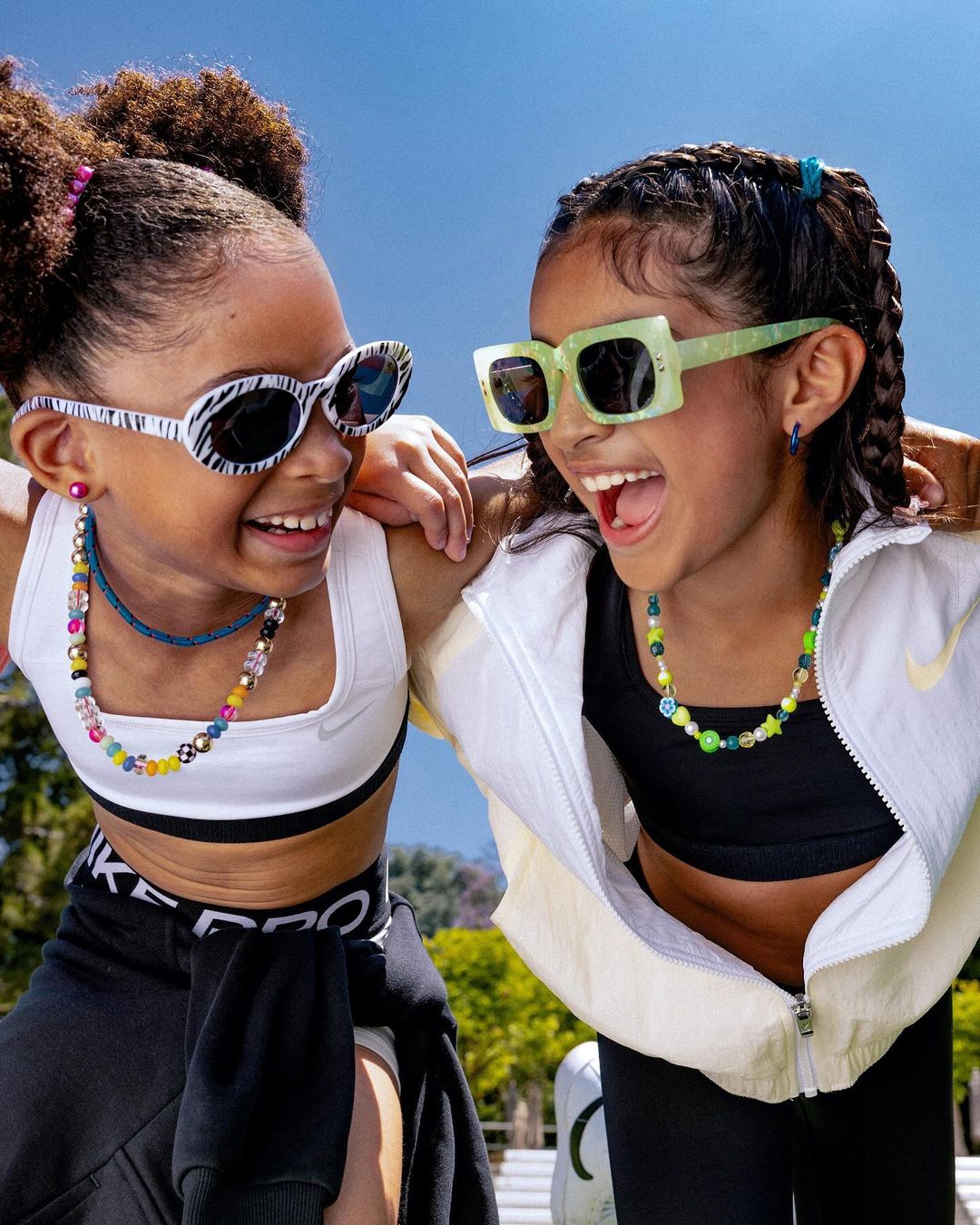 Two happy young girls smile wearing sunglasses and colorful beaded necklaces against a clear blue sky in a casual outdoor setting.