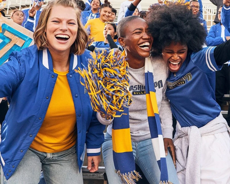 Three cheerful women, dressed in sports jackets, laugh amid a larger group of spectators at an outdoor sporting event.
