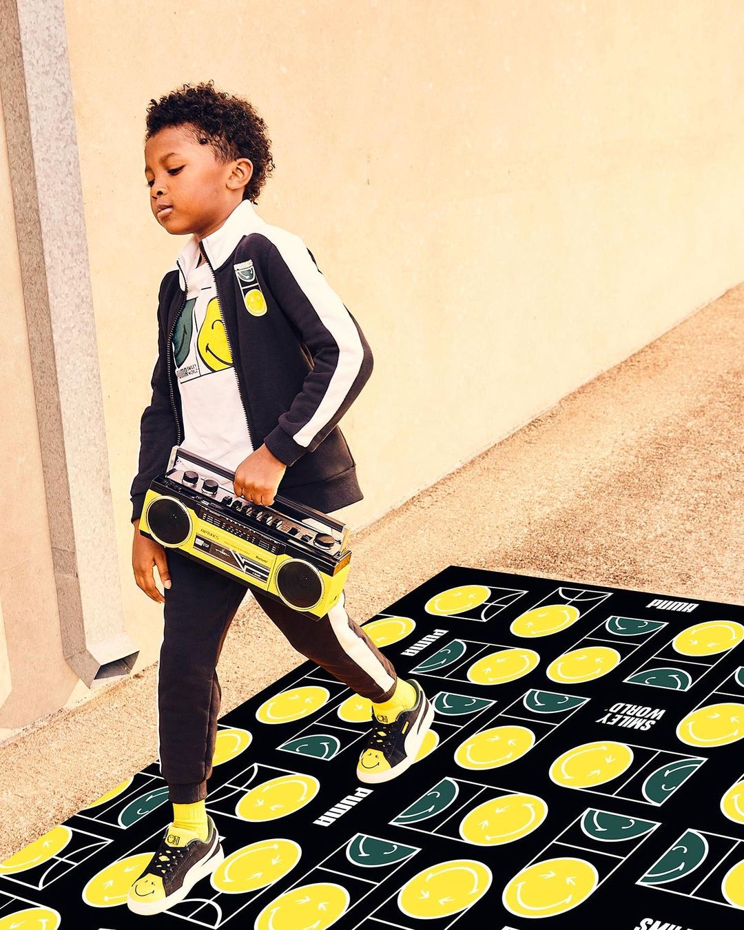 Young boy in a black and white Puma tracksuit carrying a boombox. He is standing on a Smiley World rug with yellow and green smile faces.