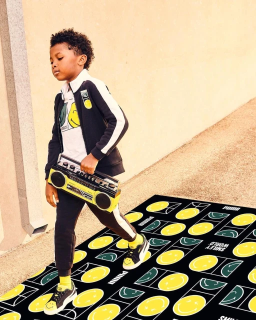 Young boy in a black and white Puma tracksuit carrying a boombox. He is standing on a Smiley World rug with yellow and green smile faces.