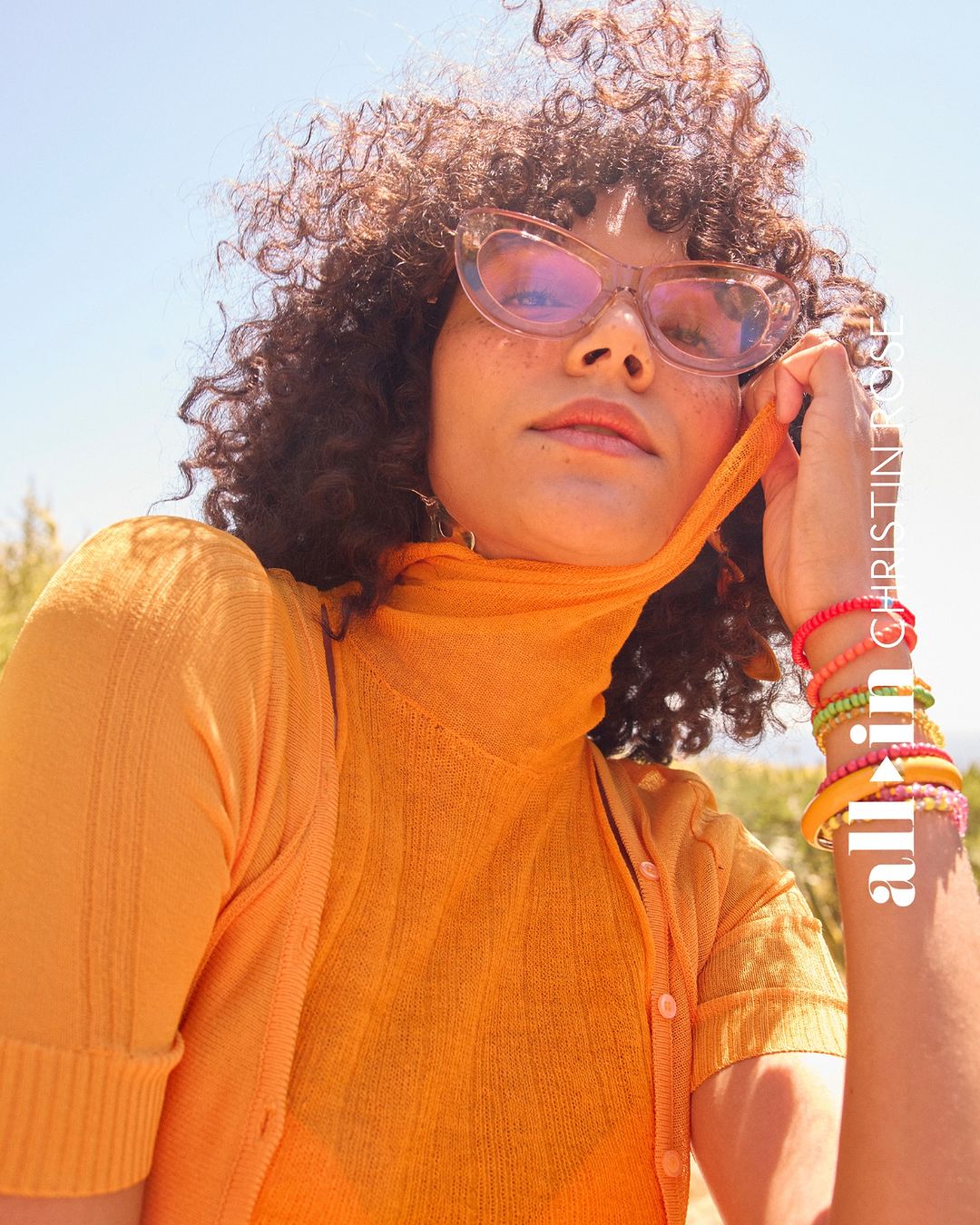 Woman with curly hair poses wearing orange turtleneck and cardigan with bracelets. Bright blue sky in background.