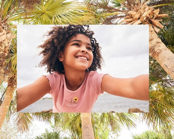 A young girl with curly hair smiling in a pink t-shirt, framed by palm trees and a body of water in the background.