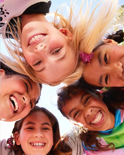 A group of five smiling children, diverse in appearance, are seen from a low angle, their faces filling the frame against a bright sky.