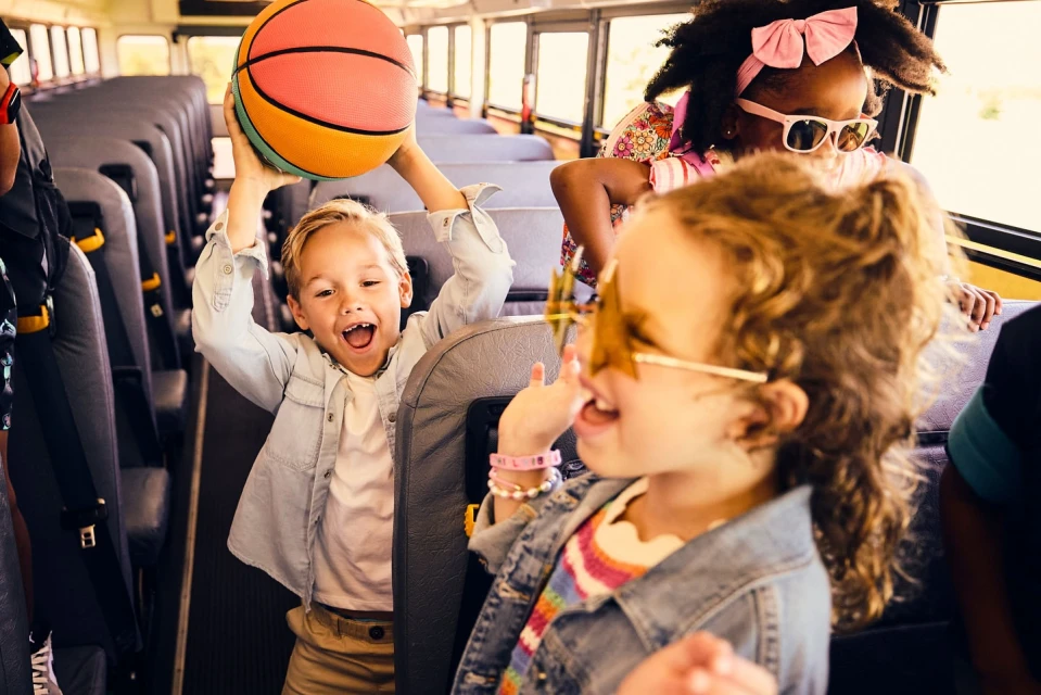 Children are having fun on a school bus with a basketball. Three kids smile as they play inside the bus.