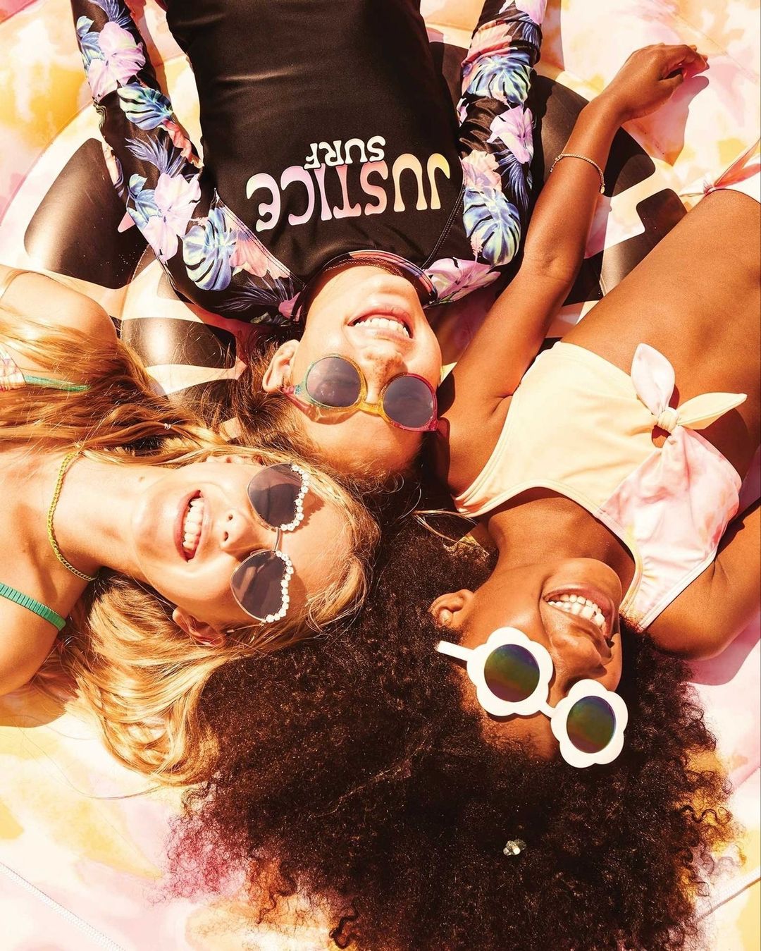 Three smiling young girls laying on a colorful inflatable pool float, wearing sunglasses and swimwear in an outdoor setting, overhead shot.