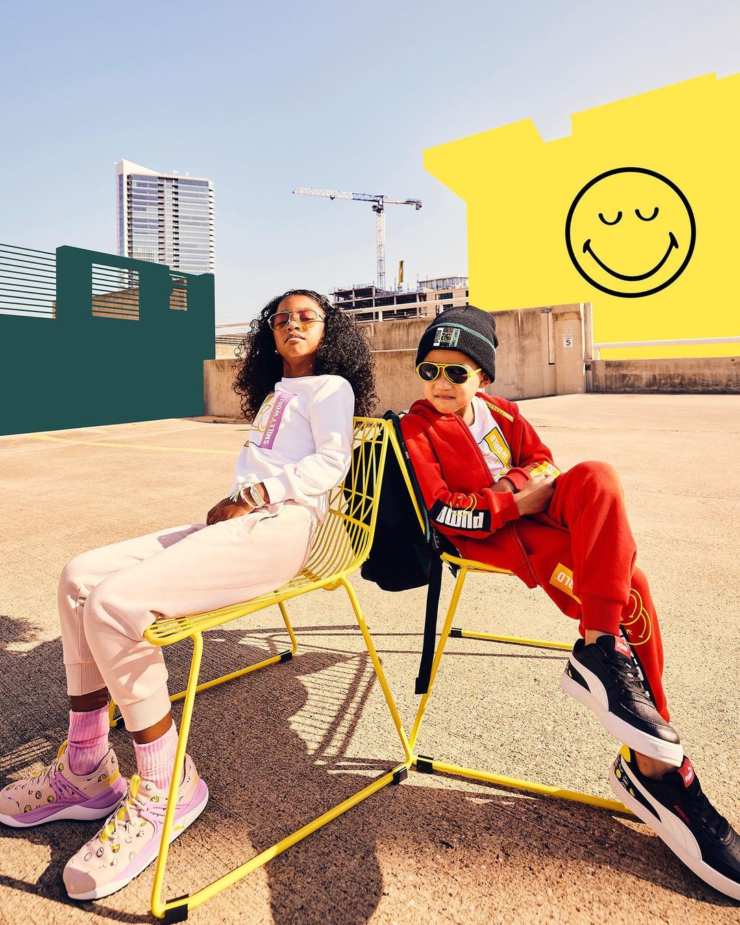 Two kids, a boy and a girl, sit in yellow chairs on a rooftop against a building backdrop, both wearing Puma tracksuits and sneakers.