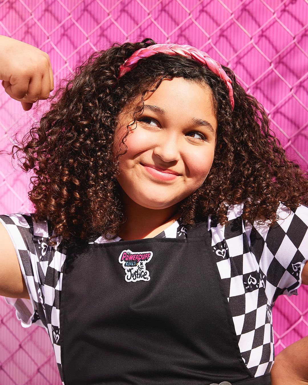 Girl with curly hair, wearing a headband and apron, poses in front of a pink chain-link fence, smiling with a flexed arm.
