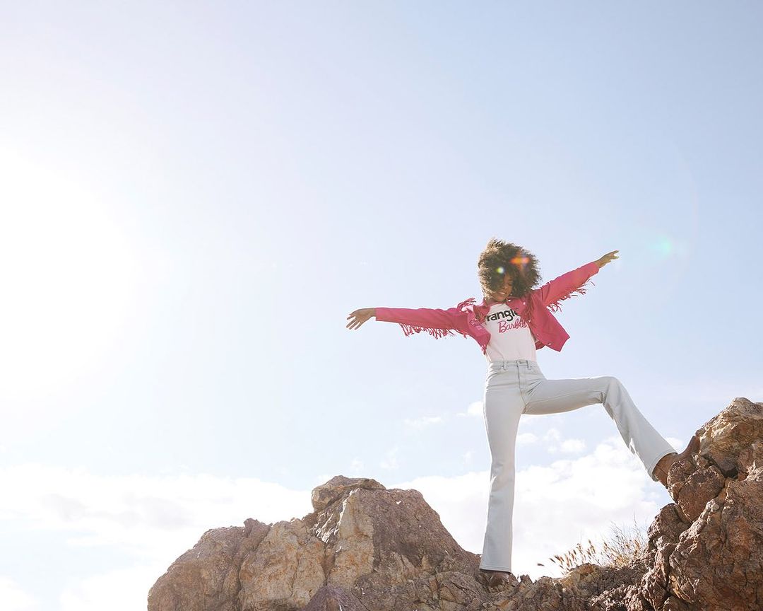 Woman in pink fringed jacket and jeans stands on rocky terrain against blue sky. Fashion-themed outdoor shot with natural light.