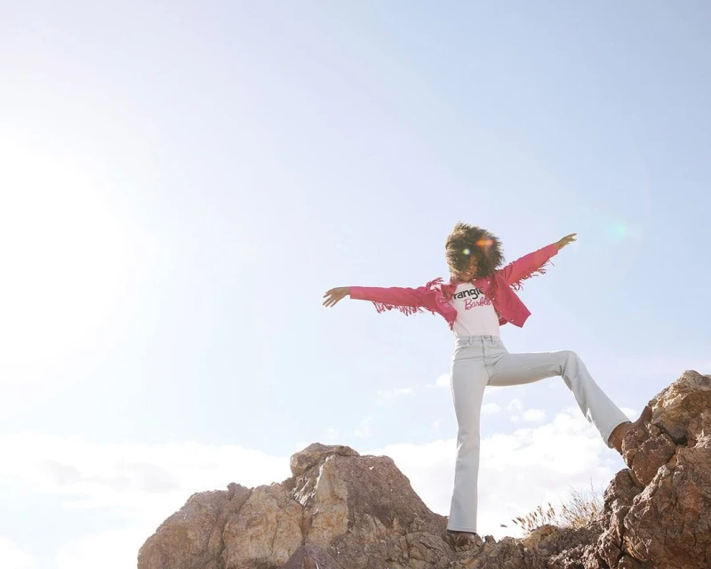 Woman in pink fringed jacket and jeans stands on rocky terrain against blue sky. Fashion-themed outdoor shot with natural light.