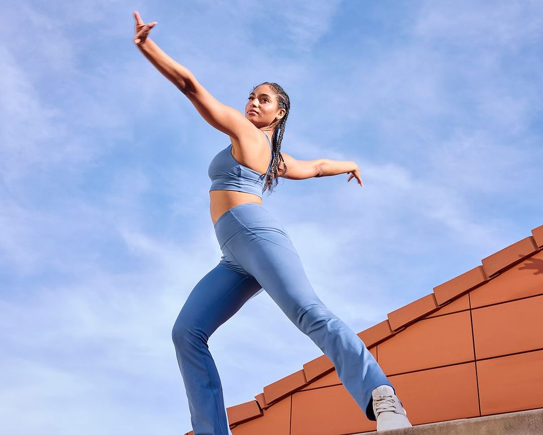 Athletic woman in blue sportswear strikes a dynamic pose on a rooftop against a bright sky, showcasing fitness and modern urban lifestyle.